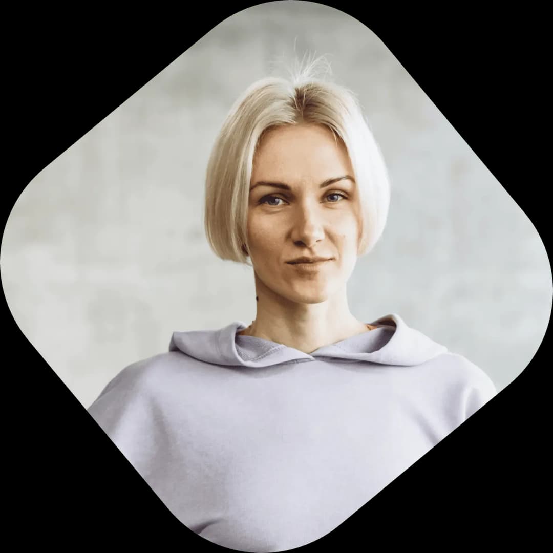 Portrait of a woman with short blonde hair wearing a light purple hoodie, against a gray blurred background.